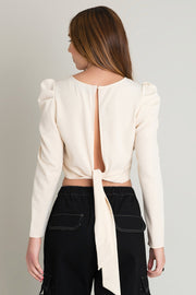Blusa de cuello redondo, manga larga de hombro abullonado y escote en espalda. Cierre posterior con botón, ojal y lazo amarrable.