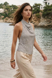 Blusa corta de estampado rayas, fit fluido, sin mangas, cuello halter fluido con plisados, escote en espalda y lazo amarrable posterior. Cierre posterior con botón y ojal.