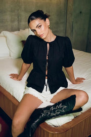 Blusa de fit amplio, cuello redondo con plisados, manga corta abullonada con plisados en hombros y rib elástico, peplum con olanes en bajo y cierre frontal con lazos amarrables.