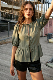 Blusa de fit amplio, cuello redondo con plisados, manga corta abullonada con plisados en hombros y rib elástico, peplum con olanes en bajo y cierre frontal con lazos amarrables.