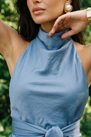 Blusa de fit recto, halter con cuello fluido, sin mangas, lazo amarrable en cintura, cierre posterior con botón y ojal.