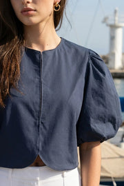 Blusa corta de fit recto, cuello redondo, manga corta amplia globo con volumen en borde, cierre frontal con broches ocultos y bajo curveado frontal.