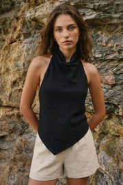 Blusa asimétrica de fit fluido, sin mangas, cuello halter fluido con plisados, escote en espalda y lazo amarrable posterior. Cierre posterior con botón y ojal. Detalle de bajo frontal asimétrico.