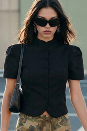 Blusa corta de fit ajustado, manga corta abullonada con plisados en hombros, cuello mock, pinzas frontales, bajo curveado frontal y cierre con hilera de botones forrados.