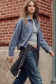 Chamarra corta de gamuza, fit oversized, cuello camisero, manga larga con puño abotonado, corte de bata frontal y posterior, bolsillos frontales de parche y cierre frontal con hilera de botones en contraste.
