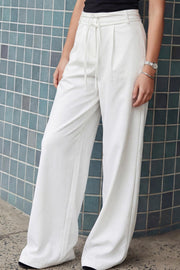 Pantalón de fit wide leg, cintura alta con pretina, trabillas y doble lazo amarrable, pinzas frontales y bolsillos delanteros. Cierre frontal con broche y cremallera ocultos.