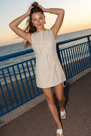 Playsuit de lino, fit ajustado, cuello barco, sin mangas, pinzas frontales, lazo amarrable posterior y cierre con cremallera oculta. Detalle frontal de lazos decorativos.