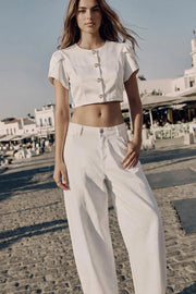 Blusa corta de fit recto, manga corta amplia con abertura, olanes y plisados en hombros, cortes frontales con pinzas, cuello redondo y cierre frontal con hilera de botones en contraste.