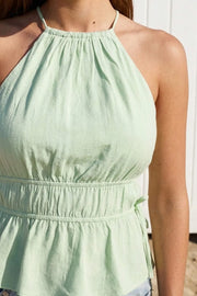 Top corto de lino, cuello halter con plisados, tirantes delgados, detalle de rib con plisados y lazos laterales en costados, peplum con olanes en bajo y cierre posterior con botón y ojal.