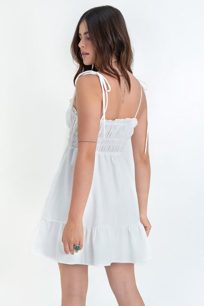 Vestido corto de escote recto con plisados, rib y olanes en borde, tirantes delgados con nudo amarrable, fruncido con rib elástico y plisados en cintura, corte en a y bajo con olanes en borde.